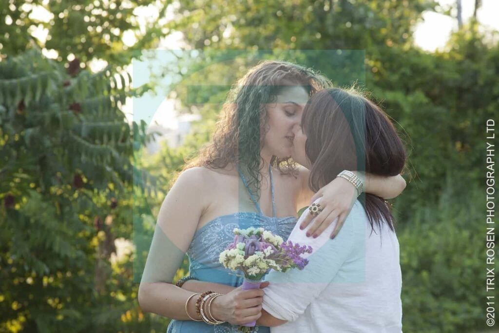 Stephanie and Kristen sharing a kiss outdoors while holding a bouquet, captured in a romantic lesbian love photograph by Trix Rosen.